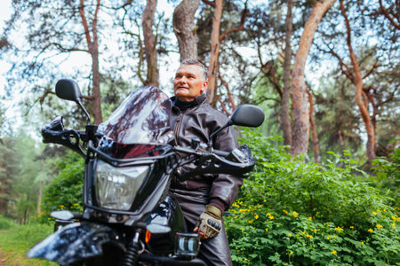 Senior motorcyclist riding motobike outdoors. Retired man in leather jacket resting after ride in summer forest. Summer road trip. Spaceの写真素材