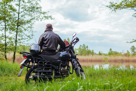 Back view of senior biker sitting on motobike outdoors. Man admiring river landscape relaxing. Rider traveling enjoying natureの写真素材