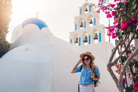 Santorini tourist with backpack walks by church with blue dome and blooming bougainvillea flowers in Akrotiri at sunset. Summer vacationの写真素材