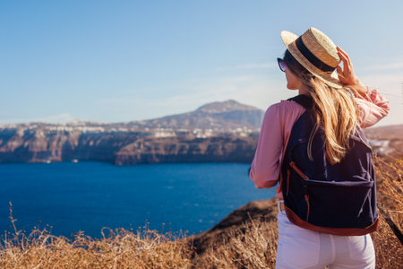 Traveler woman admires Santorini island, Greece enjoying sea landscape. Happy hiker with backpack looks at Caldera. Tourismの写真素材