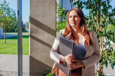 Confident worker in suit holding laptop by window. Caucasian young business woman standing in modern office by window.の写真素材