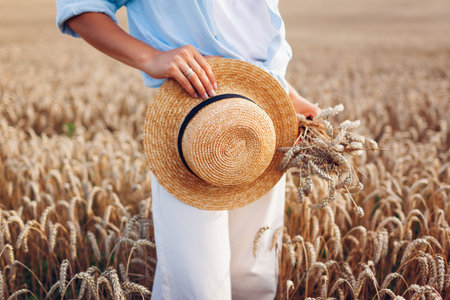 Close up of summer female accessory and clothes. Woman holding straw hat and bundle of wheat wearing linen shirt in field at sunset. Rustic boho styleの写真素材