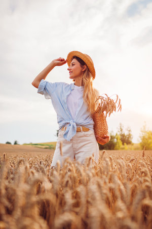 Portrait of young woman walking among wheat in summer field wearing straw hat holding handbag at sunset. Girl relaxing enjoying landscape picking wheatの写真素材