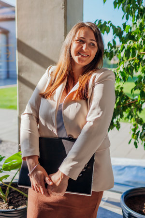 Confident entrepreneur in suit holding laptop by window. Caucasian young business woman standing in modern office by window.の写真素材