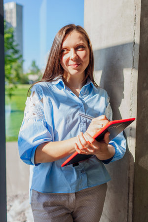 Smiling confident office worker holding tablet standing by window. Caucasian young woman using modern digital technologies. Business concept. Spaceの写真素材