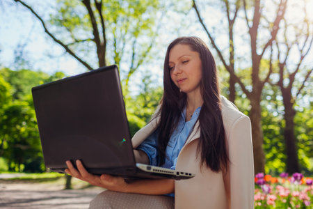 Outdoor portrait of young business woman working in spring park on laptop. Female entrepreneur uses internet in public gardenの写真素材