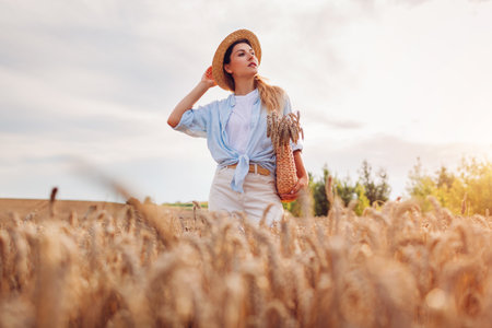 Portrait of happy young woman walking among wheat in summer field wearing straw hat holding handbag at sunset. Organic natural clothing and accessoriesの写真素材