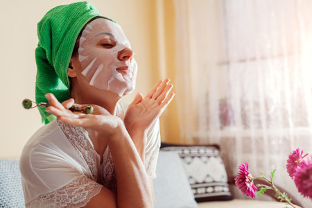 Portrait of woman with facial sheet mask applied using jade roller beauty tool and gua sha at home after bath for unti age massageの写真素材