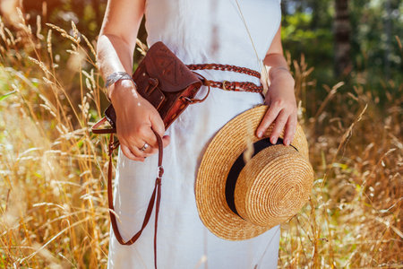 Close up of brown boho leather purse and straw hat. Stylish woman wearing white dress, jewelry, holding handbag outdoors. Fashionable female rustic boho accessoriesの写真素材