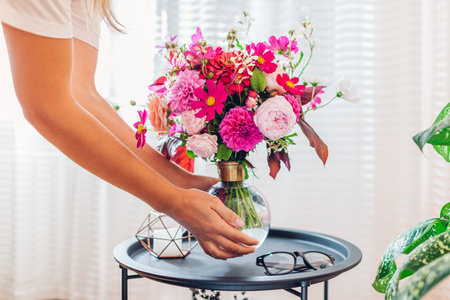 Woman puts bouquet of pink flowers in transparent vase on coffee table. Taking care of interior coziness and summer decor at home. Close upの写真素材