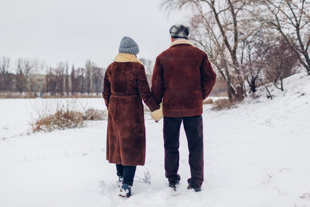 Back view of senior family couple walking outdoors during snowy winter weather. Man and woman hold hands on date.の写真素材