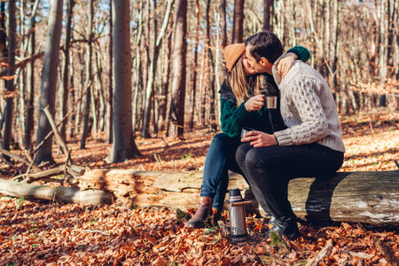 Happy loving couple kissing drinking tea from a flask relaxing in autumn forest. Man and woman sitting on trunk having coffee from vacuum flask. People enjoying date outdoorsの写真素材