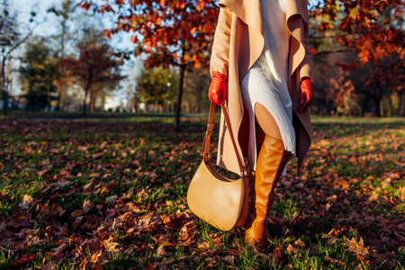 Fall female outfit. Fashionable woman wearing stylish white knitted dress, knee high boots, beige coat, gloves with handbag in park among fallen leavesの写真素材