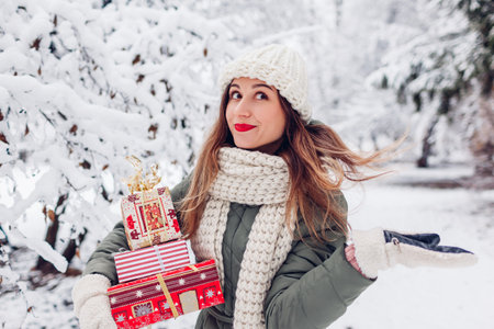 Excited young woman holding heap of Christmas presents gift boxes in snowy winter park outdoors. Festive holiday season. Girl playing with hair. Spaceの写真素材