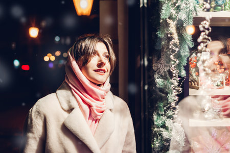 Close up of young woman looking at festive store display decorated for Christmas and New Year on city street at night. Window shoppingの写真素材