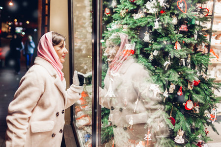 Christmas shopping. Woman looking at store showcase with decorated Christmas tree on city street at night under falling snow. Window shoppingの写真素材
