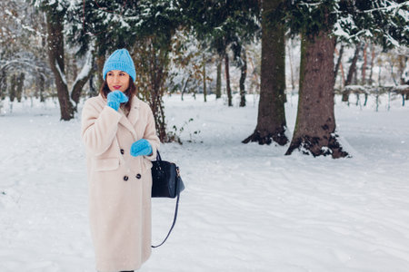 Elegant young woman walking in snowy winter park wearing white sheepskin fur coat blue knitted hat holding handbag under falling snow. Warm stylish clothesの写真素材