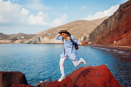 Traveler woman jumping on rock on Red beach on Santorini island, Greece. Happy girl having fun feeling extreme. Traveling and vacation conceptの写真素材