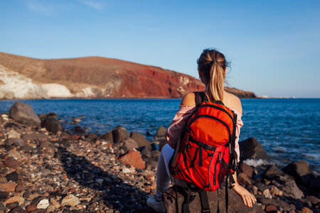 Traveler woman with backpack relaxing on Red beach on Santorini island, Greece enjoying sea and mountain landscape. Traveling and vacationの写真素材