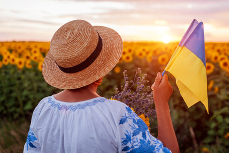 Woman patriot holds yellow and blue flags of Ukraine in sunflower summer field at sunset wearing national clothes with embroidery and hat. Independence Day of Ukraine. Back viewの写真素材