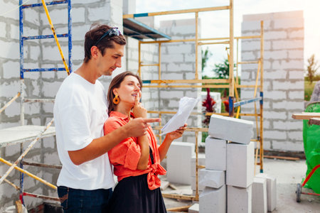 Happy young couple checking their future house under construction. Family imagines their home standing by wall made of gas block holding paper blueprint planの写真素材