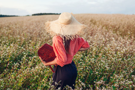 Back view of stylish woman wearing straw hat in blooming buckweat field at sunset holding leather handbag. Summer fashionable clothing and accessories. Natural organic linen shirtの写真素材