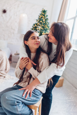 Mother hugging her daughter by decorated Christmas tree at home. Holiday winter interior. Happy family posing in festive studioの写真素材