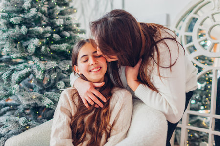 Happy mother kissing her teenager daughter at home by decorated Christmas tree. Family celebrating New Year holidayの写真素材