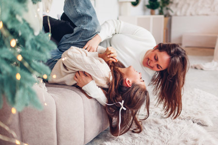 Happy family having fun at home for Christmas. Mother tickling her daughter lying upside down on sofa. Festive cozy interiorの写真素材