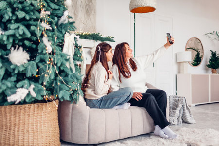 Holiday family selfie at home for Christmas. Mother and her daughter taking picture using smartphone relaxing on couch by Christmas treeの写真素材