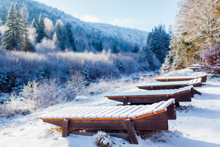 View of empty wooden chaise longue placed by mountain river side in Carpathians. Snowy winter landscape with group of deck chairs. Ski resortの写真素材