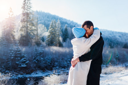 Young couple of tourists in love hugging on background of winter Carpathian mountains by snowy river enjoying landscape. Travelling on Valentines dayの写真素材