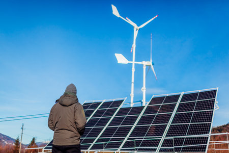 Worker checking solar energy panel and wind turbines power generator in mountains at sunset. Production of renewable alternative source of energy. Influence of mankind on environmentの写真素材