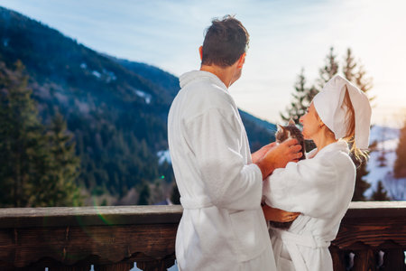 Back view of couple holding cat on hotel balcony enjoying winter mountain landscape wearing bathrobes. Travel with pet. Man and woman relaxing playing with domestic animal on terrace at homeの写真素材