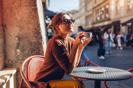 Happy stylish woman sitting in cafe outdoor drinking coffee. Woman enjoying architecture of Lviv city on street relaxing on terrace.の写真素材