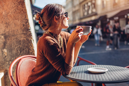 Young stylish woman sits on cafe terrace outdoor drinking coffee. Woman enjoys architecture of European city looking at streetの写真素材