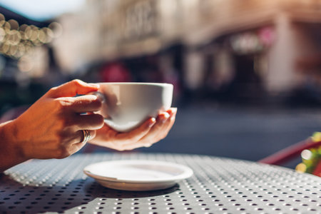 Close up of cup of coffee in female hands. Woman drinking hot beverage sitting at table in outdoor cafe at sunset. Spaceの写真素材