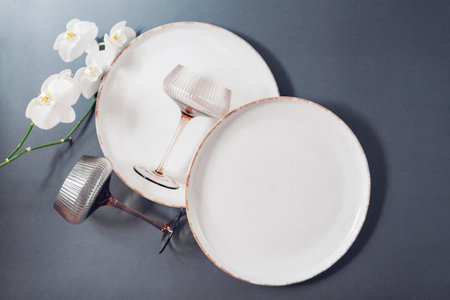 Top view of glazed ceramic dishware and cocktail glasses on grey background. Set of white speckled dinner plates decorated with flowering orchidの写真素材