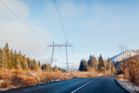Electricity pylon. High-voltage electric transmission tower surrounded with forest in mountains. Winter landscape of power lines by road. Industrial technology among natureの写真素材