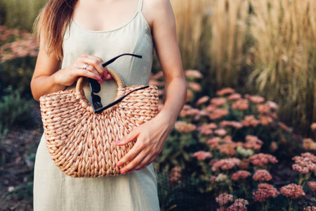 Close up of stylish organic handbag. Fashionable woman wearing summer linen dress holding wicker straw purse and sunglasses in park.の写真素材