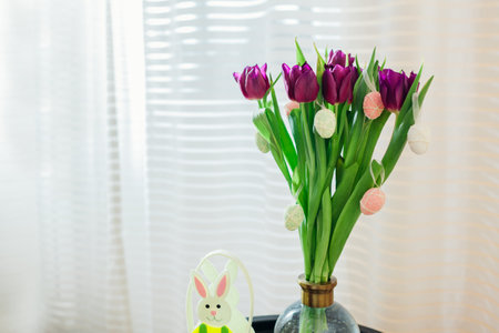 Easter home decor. Eggs hung on purple tulips in vase by bunny. Bouquet of spring flowers blooming...