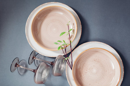 Top view of glazed ceramic kitchenware on grey background. Set of white, beige speckled dinner and soup plates with cocktail glasses decorated with spring plant branches.の写真素材