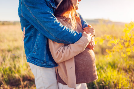 Pregnancy. Pregnant family couple walking on spring meadow at sunset. Husband hugging his wife from behind. Close upの写真素材