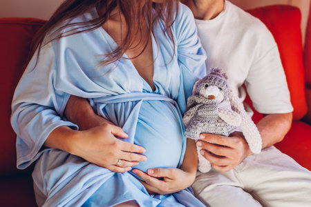 Pregnancy. Young family couple hugging at home sitting on couch holding kids toy. Future parents expecting babyの写真素材