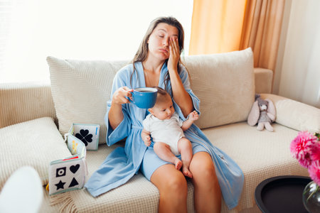Tired exhausted young mother drinking morning coffee with newborn baby sitting on her lap at home. Postpartum depressionの写真素材