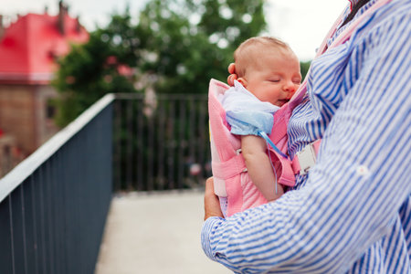 Infant girl sleeping in baby carrier. Father holds his daughter outdoors supporting child on chest. Parent-facing positionの写真素材