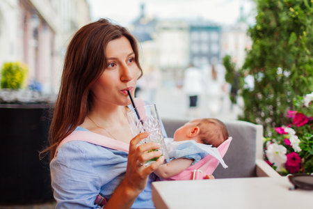 Postpartum. Young mother relaxes in outdoor cafe with baby girl sleeping in carrier. Woman drinks on summer terrace.の写真素材