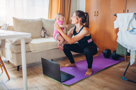 Postpartum recovery workout at home. Young mother exercising together with little baby girl on yoga mat. Squatting with infantの写真素材