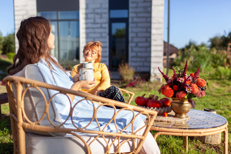 Family having coffee drinks in fall garden by house. Mother and daughter relaxing on outdoor furniture decorated for autumnの写真素材