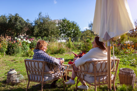 Family having coffee drinks in fall garden. Mother and daughter relaxing on outdoor furniture covered with blankets. Breakfast on terrace with autumn decor. Back viewの写真素材